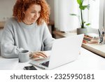 Portrait of curly haired redhead caucasian female in wired headphones spending leisure time using laptop during coffee break, sitting at kitchen table with smartphone, visiting popular web pages