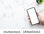 Hand holding smartphone with white blank mock up screen over white marble office desk table with supplies. Top view image.