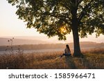 Attractive young girl in casual clothes sitting under big tree with book and looking at the beautiful sunset, autumn sunny day