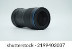 Small photo of A black manual 65mm macro photography prime lens for APSC cameras, with blue accents. Lying on a white surface in front of a white background, casting shadows on the surface.