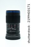 Small photo of A black manual 65mm macro photography prime lens for APSC cameras with blue accents, standing upright on a white surface, isolated on white background. Without lens cap attached.