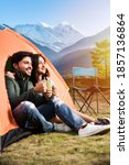 Happy young asian indian couple camping, sitting by tent at campsite relaxing and drinking tea.