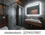 Small photo of Black bathroom with gold trim. Corner bathroom, chic mirror with washbasin. Lamps trimmed with gold. Bathrobe and towels on a hanger. White floor mat.