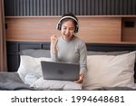 Young asian woman with headphone showing yes gesture with hand after receive email good news while sitting to working and listening to music from laptop on the bed in home bedroom