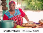 Small photo of Portrait of an adult woman standing in her kitchen looking at a camera. Proud African American woman in colorful Garifuna dress.
