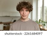 Close up of a young handsome positive curly man with blue eyes with a smiling face wearing casual beige t-shirt looking at camera and standing alone in morning in the modern kitchen at home.