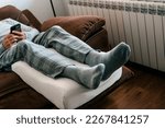 Small photo of a man leans his legs on a leg elevation pillow, made of memory foam, while is resting on the sofa