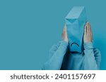 closeup of a man with a blue paper bag in his head with a sad mouth drawn in it, and his hands around it, on a blue background with some blank space on the left