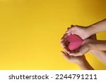 Hands of two people holding a red heart shape on yellow background with copy space. 