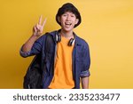 Cheerful male college student showing two fingers v-sign peace greeting gesture in casual clothes, headphones around his neck and wear a bucket hat 