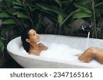 Young woman blowing soap bubbles and having fun while lying in a bathtub full of foam. Tropical home spa concept. Charming lady relaxing in the bath and enjoying a beauty and skincare day