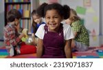 Small photo of Close up portrait of smiling little African-American girl looking at camera at primary school. Adorable preschool kid sitting in playroom of kindergarten