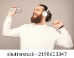 Relaxed young man wearing white sweatshirt, headphones and holding phone enjoys good music. Studio shot over grey background.