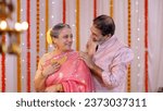Small photo of A charming old man giving a Diwali gift to his beautiful wife - putting necklace, old age love, celebrating golden jubilee, happy anniversary, gold necklace. Old couple celebrating Diwali festival ...