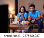 Small photo of Friends in Indian jerseys sitting on the couch and watching a live cricket match on TV television - cricket fever. Indian cricket fan wearing an Indian jersey - excited, cheering for a team