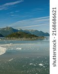 Small photo of Disenchantment Bay, Alaska, USA - July 21, 2011: Portrait, Floating ice pieces on blueish water in front of green forested mountains and islet under blue cloudscape.