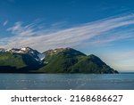 Small photo of Disenchantment Bay, Alaska, USA - July 21, 2011: Green forested and snow covered mountain group under blue cloudscape block partly exit to ocean. Blue water with floating ice pieces