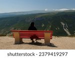 A Muslim woman in a black niqab sits on a bench against the backdrop of mountains. Horizontal photo