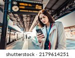 Young woman with departure times behind her waiting for her train while holding her mobile phone - Woman looking at the clock in the train station while her train is delayed - Transportation concept