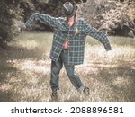 Small photo of A lady with a hat on her face and a plaid shirt with overly long sleeves stands in a pose on a green lawn. Parody of a garden scarecrow