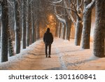 Young man in black clothes alone slowly walking on snow covered sidewalk through alley of trees. Peaceful atmosphere in snowy winter night. Back view.