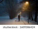 Young woman alone slowly walking on sidewalk under yellow street lights in mist. Foggy air. Peaceful atmosphere in snowy night. Back view.