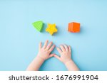 Baby hands playing with green triangle, yellow star and orange square shapes on light blue table background. Pastel color. Closeup. Toys of development for little kids.