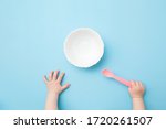 Baby hands holding pink plastic spoon and waiting food. Empty white bowl on light blue table background. Pastel color. Closeup. Point of view shot.