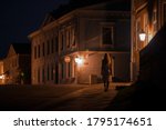 Young alone woman in white dress slowly walking on sidewalk under street lights at old town in dark summer night. Peaceful atmosphere. Spending time alone. City life. Back view. 