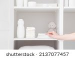 Small photo of Young adult mother hand taking white towel from inside wardrobe. Bottle of talcum powder, stack of new nappies and teddy bear on shelf. Preparing for baby diaper changing. Hygiene goods. Closeup.