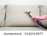 Woman hand in rubber protective glove using spray nozzle of professional vacuum cleaner and washing light beige back pillow of sofa at home. Extraction method. Commercial cleaning service. Closeup.
