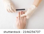 Small photo of Manicurist hand in rubber protective gloves using nail file and filing young adult man nails on white towel background. Manicure, pedicure beauty salon concept. Closeup. Care about fingernails.