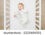 Adorable happy baby boy in white bodysuit hugging teddy bear and  lying down on back on mattress in crib at home room. Top view. 6 months old infant playing with first friend. Closeup. 