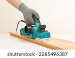 Small photo of Young adult man hands using electric jointer and shaving old wooden plank for furniture or floor on white table at light gray wall. Closeup. Preparing material for repair work of home. Side view.