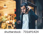Hispanic young attractive man stands in dark street in front of shop, changes songs and tracks on smartphone, listens to music in wireless headphones. Hipster with slight beard