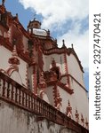 Small photo of The facade of an ancient church with white walls and red details. A cross at the top. Santa Prisca church in the touristic magical town of Taxco, in Guerrero, Mexico.