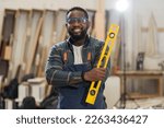 Small photo of Portrait of African American male carpenter holding precision level making new furniture at wood workshop. Portrait of joiner male working with precision level in wood factory