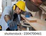 Small photo of Male carpenter using electrical circular saw electric during working in wood workshop. Male joiner wearing safety uniform, gloves, helmet and working in furniture workshop