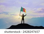 Small photo of Cameroon flag being waved by a man celebrating success at the top of a mountain against sunset or sunrise. Cameroon flag for Independence Day.