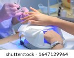 Manicurist master is covering painting client's nails top coat shellac, hands closeup. One hand drying in UV lamp. Professional manicure with gel polish in beauty salon. Beauty industry concept.