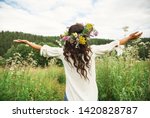 Summer lifestyle portrait of beautiful young brunnete woman with perfect long curly hair in a wreath of wild flowers. Standing back in the flower field, hands to the side. Romantic mood. Nature lover 
