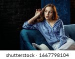Fashion portrait of beautiful elegant woman wearing stylish light blue silk blouse, white high waist trousers, sitting on sofa, posing against dark background. Copy, empty space for text