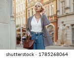 Street fashion photo of elegant  woman wearing trendy white blouse, stylish checkered blazer, high waist jeans, holding brown faux croco leather textured bag. Model walking in street of European city
