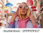 Fashionable confident girl wearing trendy pink sunglasses with yellow plastic chain, hood, blue trench coat, posing in European city. Streetstyle, street fashion conception. Close up portrait