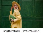 Joyful smiling woman wearing yellow hat, classic shirt, holding quilted faux leather green bag with chain, posing on green background in street. Copy, empty space for text