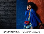Fashion portrait of happy smiling African American woman wearing  classic blue blazer, satin floral print skirt, with red textured reptile bag. Model posing near colorful walls. Copy, empty space