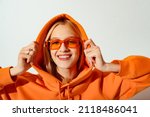 Small photo of Happy smiling fashionable woman wearing trendy orange color sunglasses, hoodie posing on white background. Close up studio portrait. Copy, empty space for text