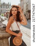 Small photo of Elegant curly brunette woman wearing trendy summer outfit with round wicker shoulder rotan bag, white top, polka dot skirt, posing in street of European city. Fashion, lifestyle concept