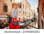 Small photo of Happy smiling fashionable woman wearing straw hat, polka dot blouse, red skirt, holding handbag, posing on the bridge in Venice. Fashion, travel, lifestyle conception. Copy, empty space for text