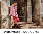 Small photo of Fashionable happy smiling blonde woman wearing stylish red beret, classic pink suit blazer, checkered midi skirt, ankle boots, posing in street of European city. Copy, empty space for text